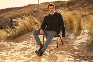 Man in black denim jacket and grey pants sitting on chair in sandy dunes, fall winter fashion