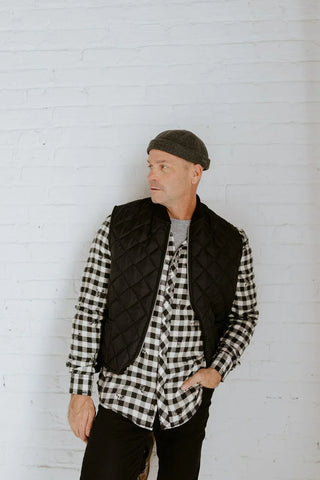 Man wearing a dark beanie, black quilted vest, and checkered shirt against a white brick wall