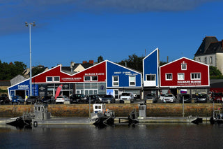Colorful waterfront buildings with marine clothing shops and parked cars in front of a harbor.