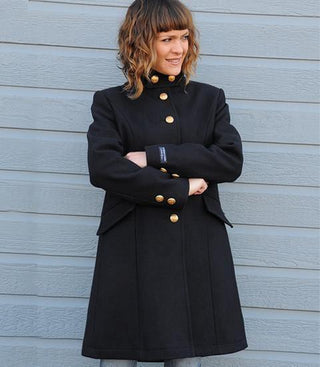 Woman in long navy wool coat with gold buttons standing against blue wall