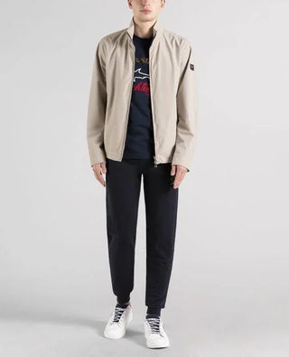 Man wearing a beige nylon zip-up jacket over a navy shirt and black pants with white sneakers