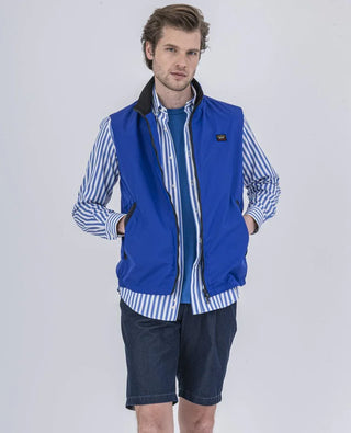 Man wearing blue nylon vest, striped shirt, and denim shorts against a white background